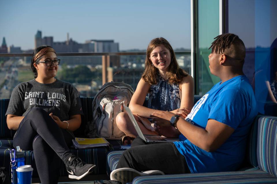 Three students chat in a sunny spot in a common area in Grand Hall.
