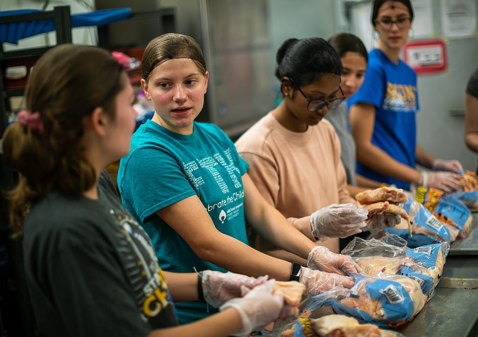 Students wearing hairnets and gloves work in a line at a kitchen counter.