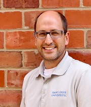 Bobby Wassel, headshot, wearing a grey SLU polo shirt, stands in front of a brick wall and smiles.