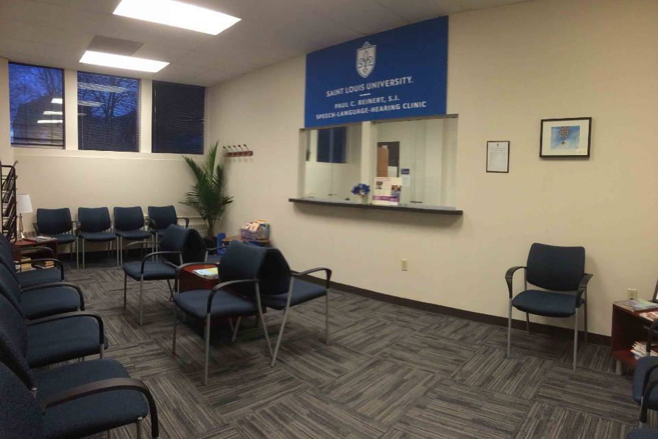Interior view of a waiting room with chairs and plants. A large sign with the SLU logo and the words "Paul C. Reinert Speech Language and Hearing Clinic" hangs above the reception window.