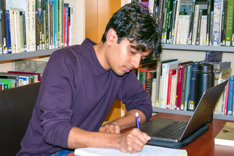 A student sitting at a table with a laptop computer while writing in a notebook.