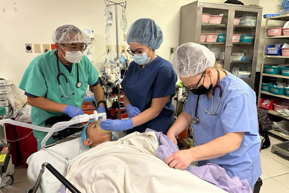 Three doctors wearing scrubs, and surgical protective equipment attend to a patient in a medical facility.