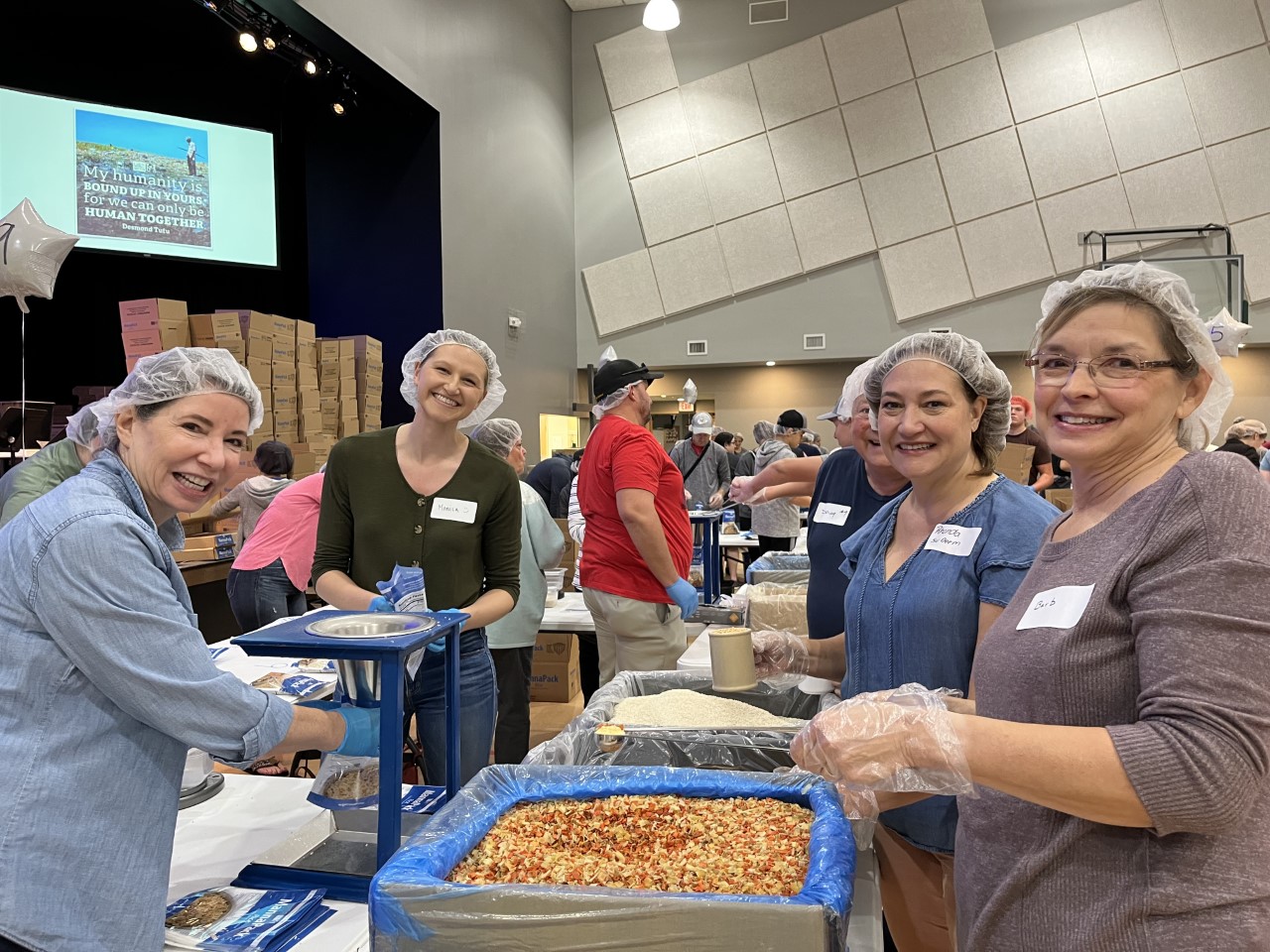 Volunteers wearing hairnets and gloves stand around a counter packing items for meals.