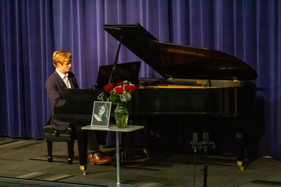A student plays at a grand piano with a table containing a vase of roses and a photo at the front.
