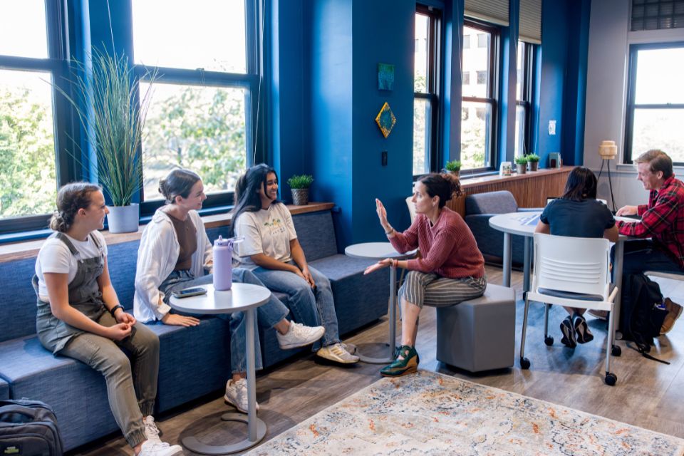 Students sit along a cushioned wall bench while talking with a faculty member.