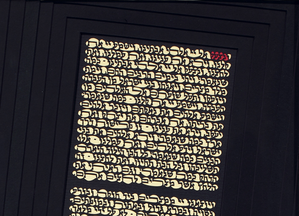 Detail of Page 13 of The Papercut Haggadah by artist Archie Granot. A tall cream-colored rectangle made up of three blocks of Hebrew text is set, leaning diagonally to the right, against a deep black background. One word at the upper right corner is set in red.