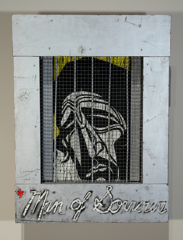 An artwork by Adrian Kellard titled Man of Sorrows. A portrait of Jesus, carved in wood and painted white against a yellow background, is seen inside a wooden frame behine chicken wire and rods, so that he appears to be in prison. The words "Man of Sorrows" are carved along the bottom edge of the frame, with a small red cross to the left.