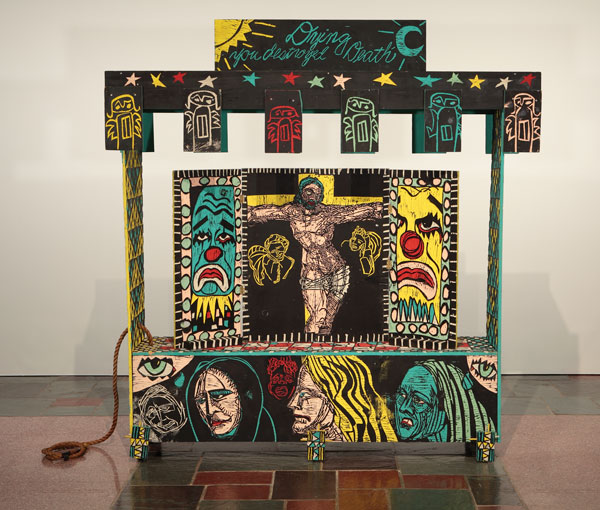 An artwork by Adrian Kellard titled Shrine. A large wagon made of carved wood panels painted in yellow, teal, red, and black, features carved drawings including Jesus on the Cross, clown faces, whimsical angels, and the words, "Dying you destroyed Death."