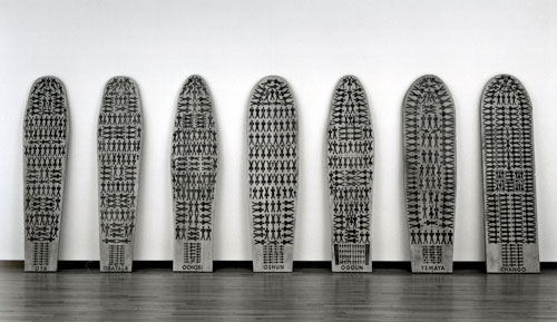 An artwork by Maria Magdalena Campos-Pons titled, The Seven Powers Come By the Sea. Seven ironing boards lean upright against a wall. Each bears a reproduction of a diagram of slaves packed into the hold of a slaving ship.