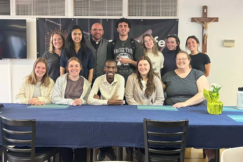 Students and a priest pose for a photo while standing and sitting behind a table.