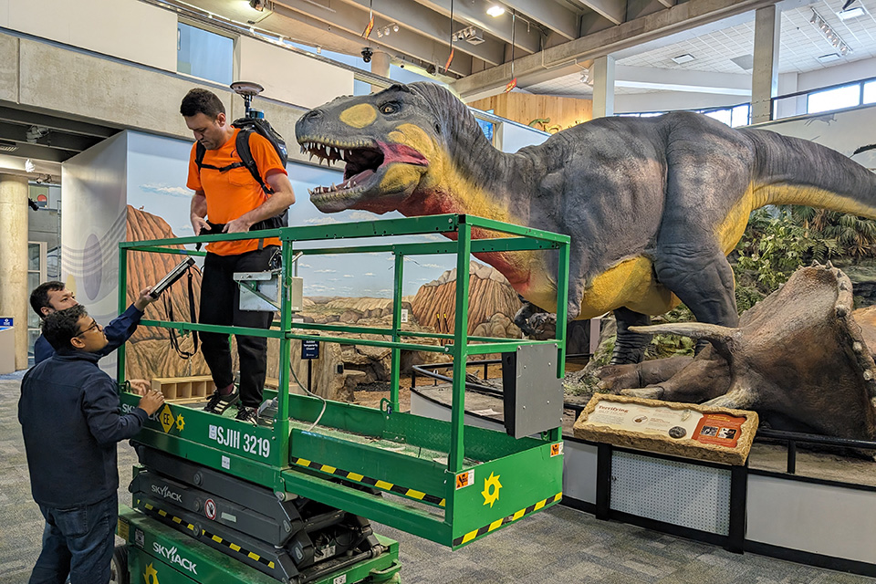 A student stands on a lift with a large piece of equipment on his back in front of one of the latex dinosaurs.
