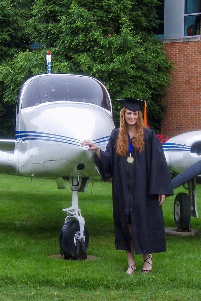 Sullivan stands near the plane in front of Parks Hall in her graduation robe.