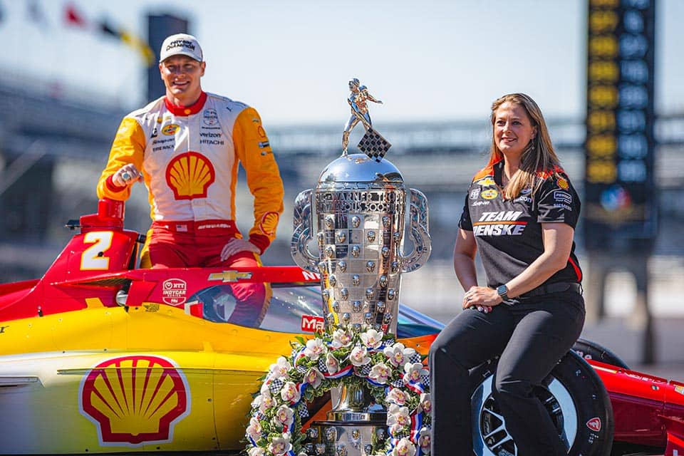 Sullivan sits in front of a racecar near a trophy won at the Indy 500 across from a driver.