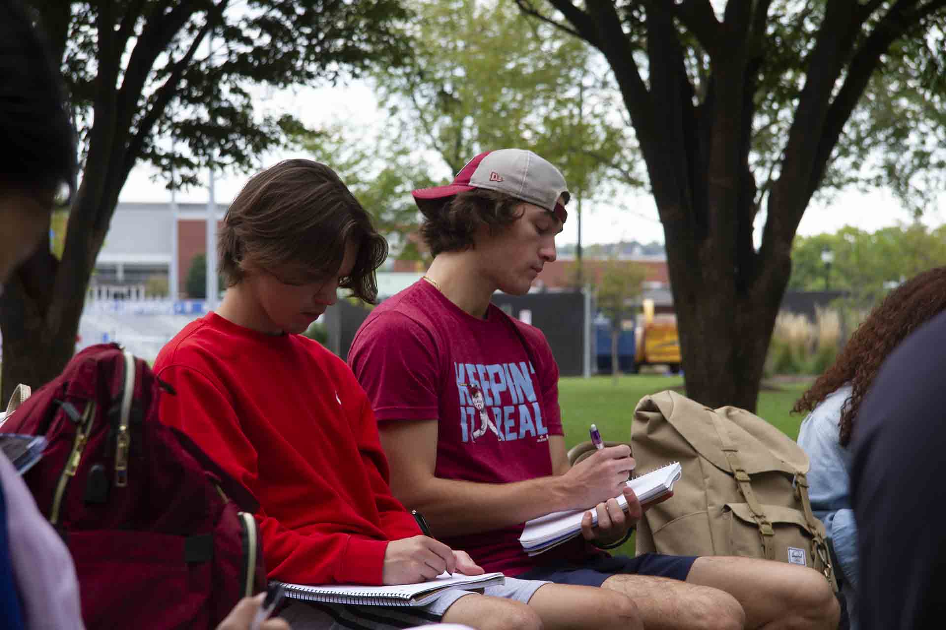 Students take notes during a class session under a tree.