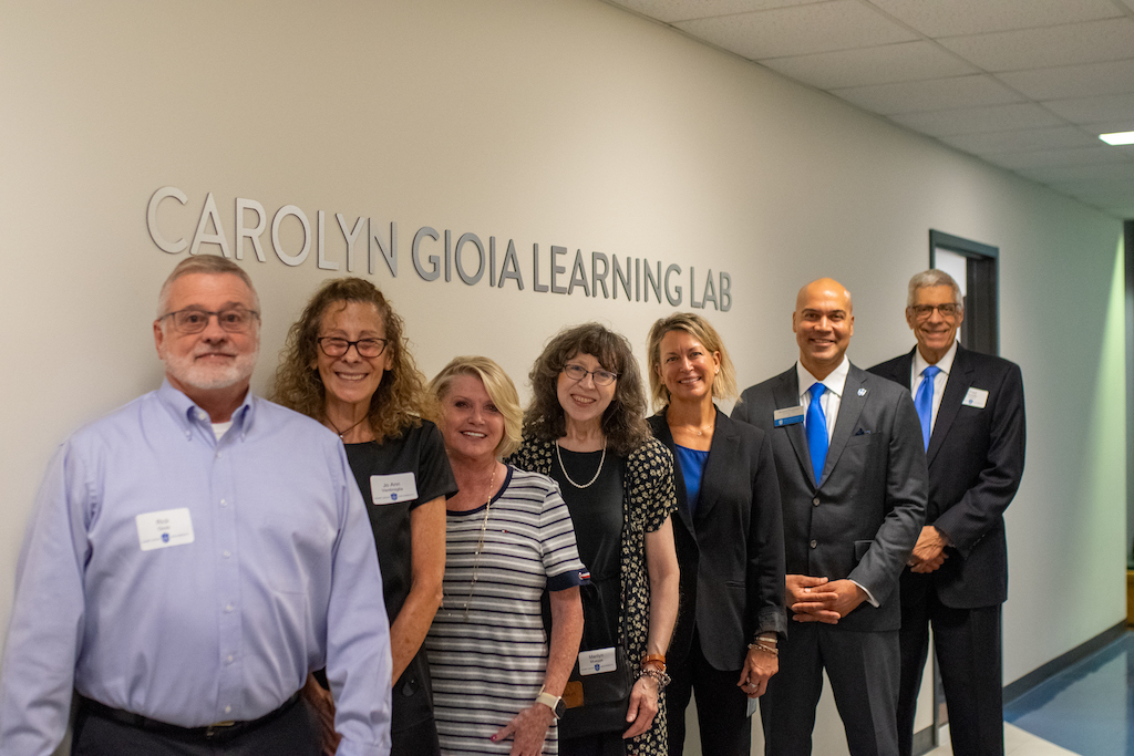 Seven people stand in front of the "Carolyn Gioia Learning Lab" wall sign inside the Allied Health Building on SLU's South Campus. 