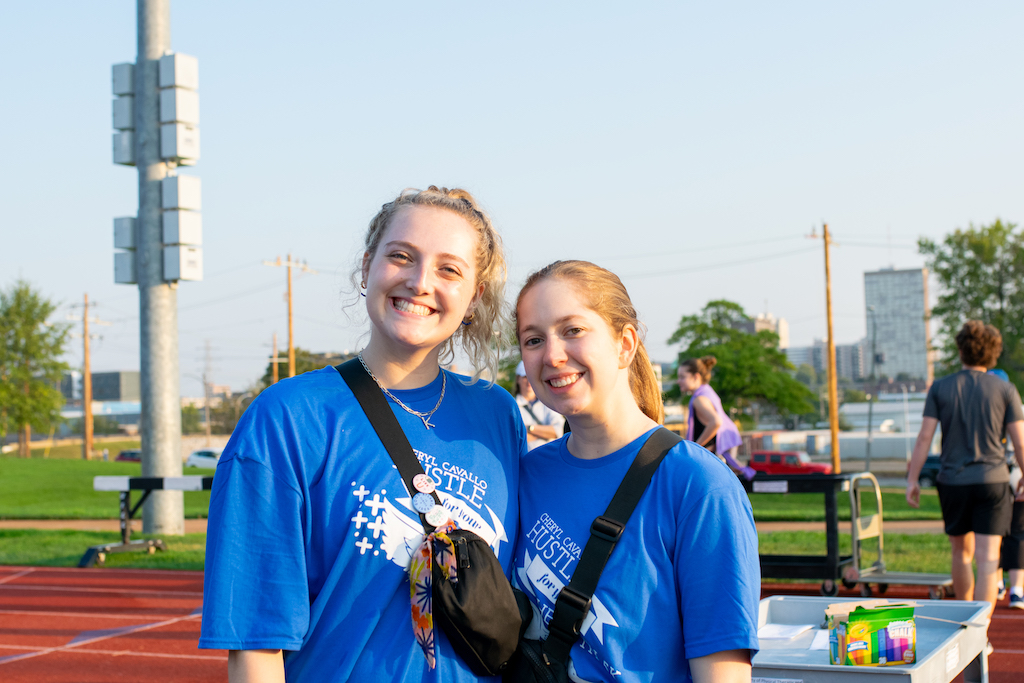 Two girls stand together outside wearing blue "Hustle for Your Health" t-shirts. 