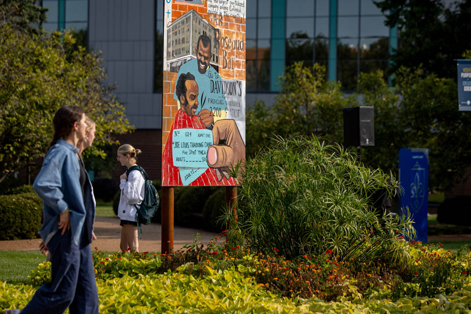 People walk past a piece from “The Legacy of Mill Creek,” an art installation by Cbabi Bayoc surrounding the fountain at Grand and Lindell Blvds. The installation honors the vibrant historically Black community that lived in the former Mill Creek Valley neighborhood. Photo by Sarah Conroy. 