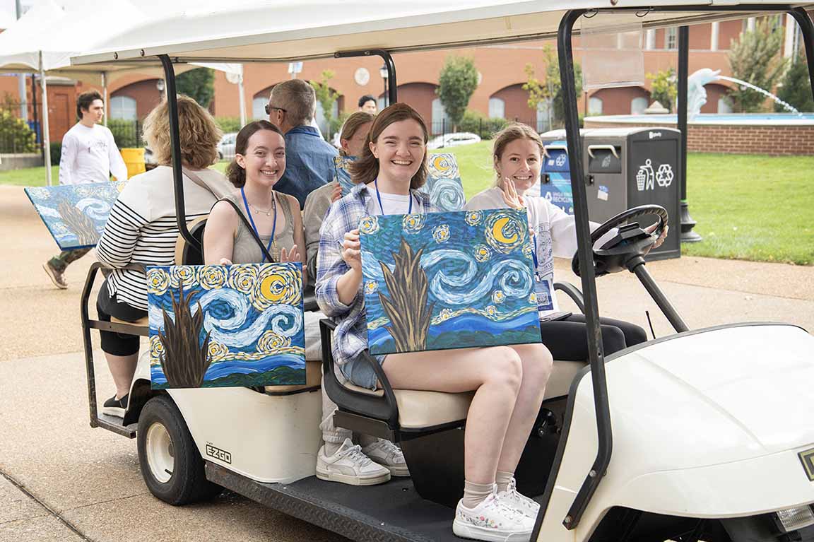 A golf car holding family members showing off artwork from the 2024 Homecoming and Family Weekend Paint and Sip drives down West Pine Mall. 