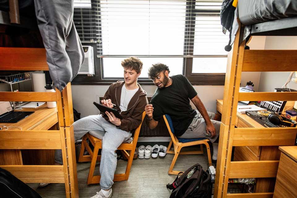 Two male students lean back in chairs to chat in their residence hall room.