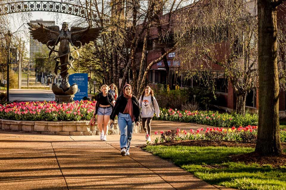 Female students walk on a path up from a parking garage surrounded by trees and tulips.
