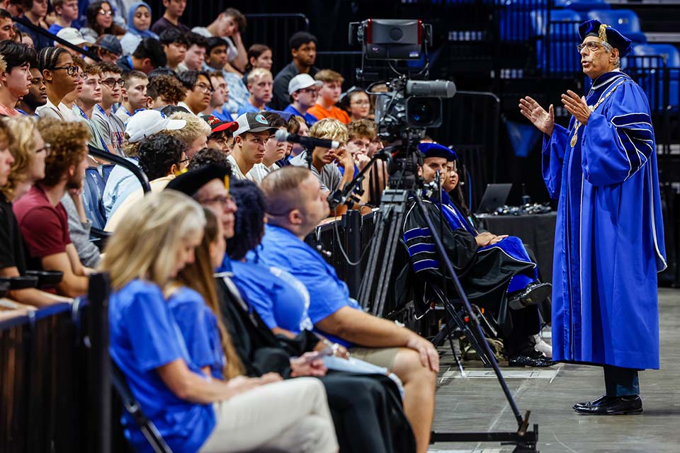 SLU President Fred Pestello wearing academic regalia addressing students in arena seating at convocation