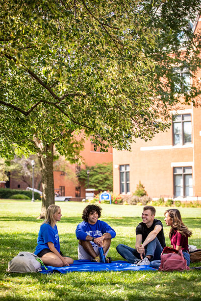 A group of four students sitting on a picnic blanket in a grassy area of SLU's campus