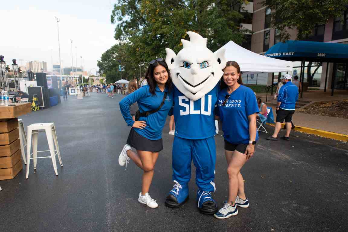 Two people in SLU spiritwear pose for a picture with the Billiken mascot in early evening during homecoming festivities. 