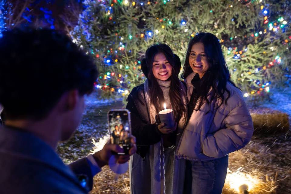 Two female SLU students bundled in winter coats pose for a photo in front of the University Christmas tree, which is covered in colored lights. One student holds a mug with a lit candle inside. Another student stands in the foreground and takes their picture on a smartphone.