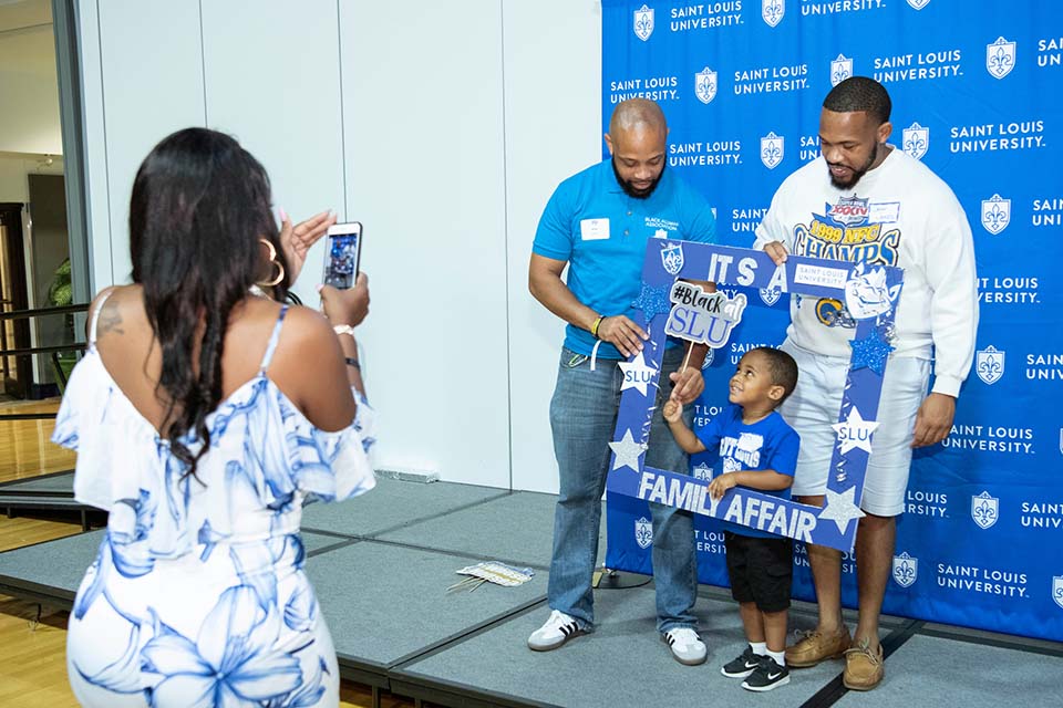 Two men and a young boy stand in front of a blue Saint Louis University background and pose for a picture holding an oversized blue photo frame adorned with stars and SLU decorations. A woman stands in the foreground and takes their photo on a cellphone.