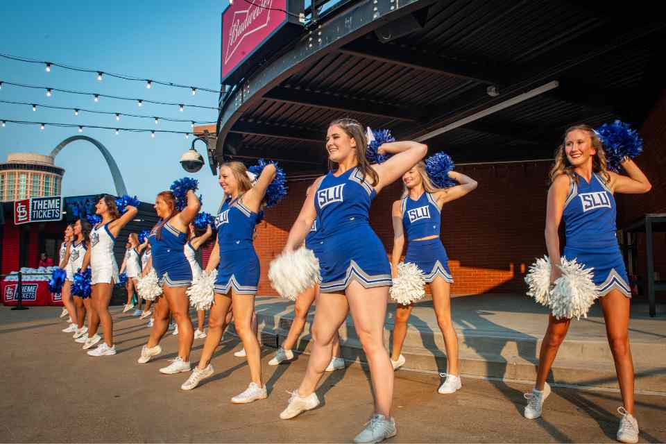 SLU's cheer team in uniform with poms in front of Busch Stadium with the Gateway Arch in the background.