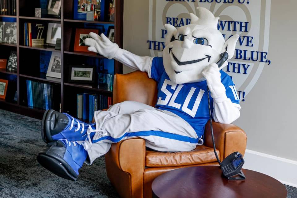 The SLU Billiken sits in a brown leather chair with his legs draped over one arm, holding a landline phone to his ear. In the background there is a bookshelf filled with many blue books and framed photographs, as well as a wall adorned with the SLU fleur de lis and the lyrics to the University's Varsity Song: "Bear we with pride and love Thy White and Blue, Sweet are thy memories, Saint Louis U!"