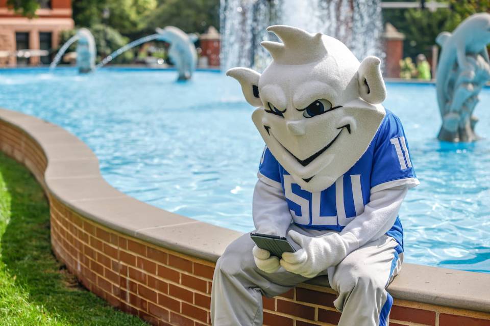 The SLU Billiken sits on the edge of the Dolphin Pond fountain on the University's North Campus while texting on a smartphone.