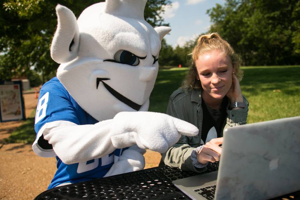 The Billiken working on a laptop in the quad with a student.