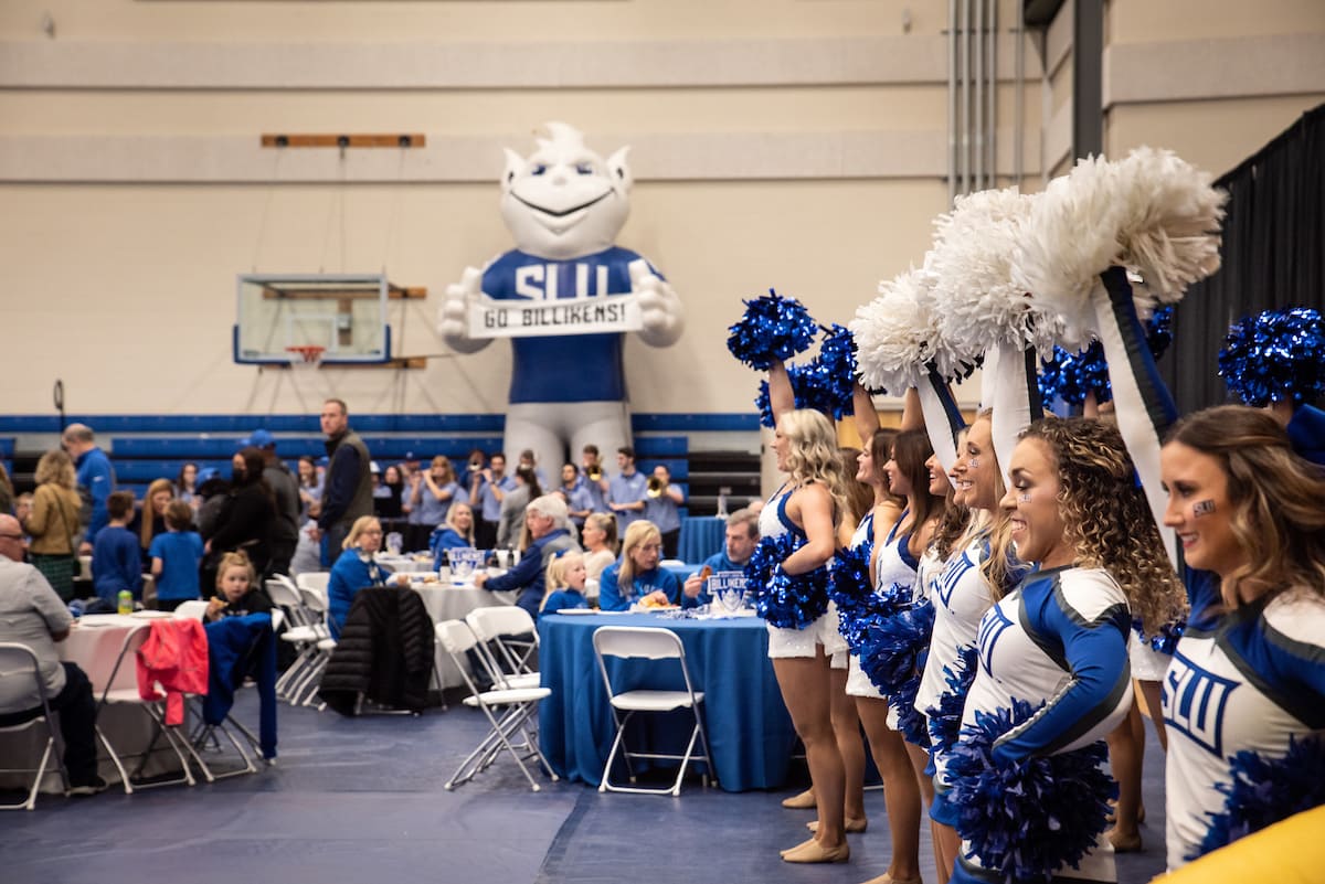Members of the SLU cheer and dance team stand in a line and smile, holding blue and white pom poms in the air. To the left, True Blue Fan Fest visitors wearing blue and white SLU spiritwear sit around tables adorned with "Saint Louis Billikens" signs. In the background, members of the SLU pep band ready their instruments in front of a basketball hoop and a large inflatable Billiken holding a sign that reads "Go Billikens!"