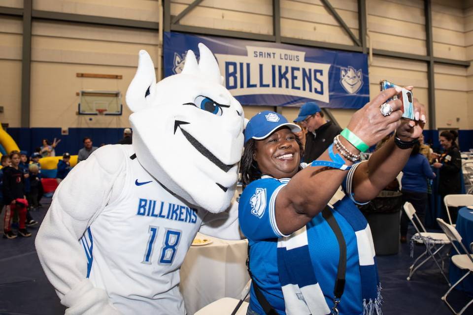 The SLU Billiken wears a white basketball jersey and poses for a selfie with a woman decked out in blue SLU spiritwear. In the background, young children play near an inflatable slide and a blue banner reading "Saint Louis Billikens" hangs on the wall. 