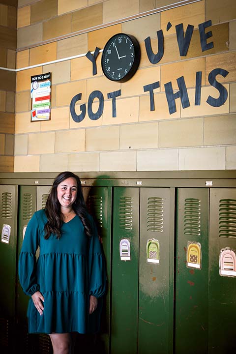 Megan Kelly, wearing a blue dress, stands in front of a row of dark green lockers in an elementary school hallway.
