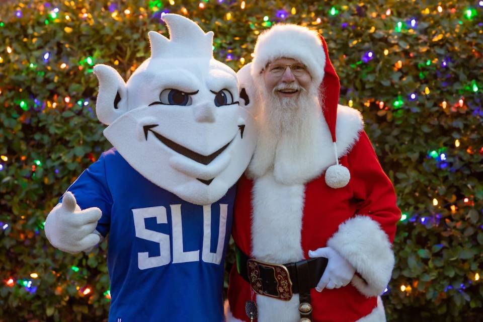 Santa Claus and the SLU Billiken pose for a photo in front of a Christmas tree adorned with colored lights.