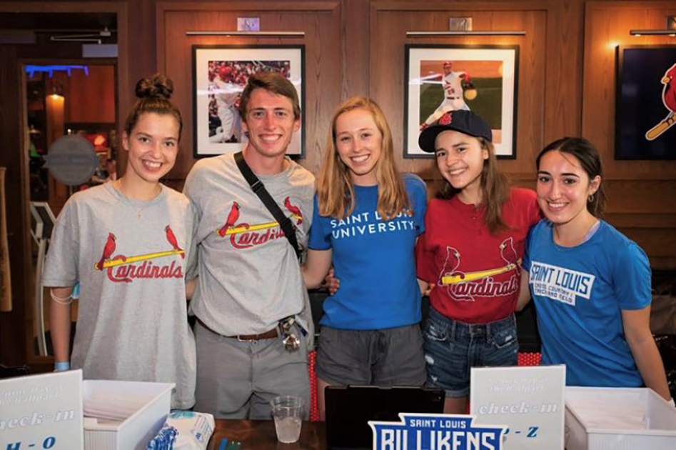 A group of five students wearing SLU and St. Louis Cardinals T-shirts pose in a wood-paneled room with framed photos of Cardinals baseball players on the wall.