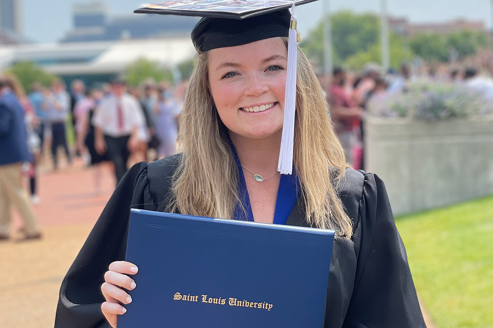 Becca Cary poses for a photo wearing a graduation cap and gown outside on campus while holding a diploma.