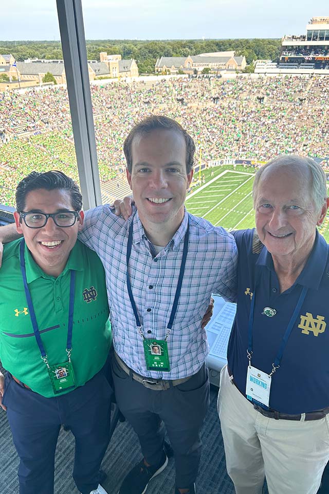 Chris Ackels Three men pose for a photo with a stadium behind them. Two of the men are wearing Notre Dame shirts.