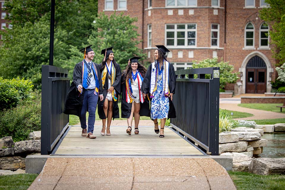 Four students walk across a bridge on campus while wearing graduation gowns and caps.