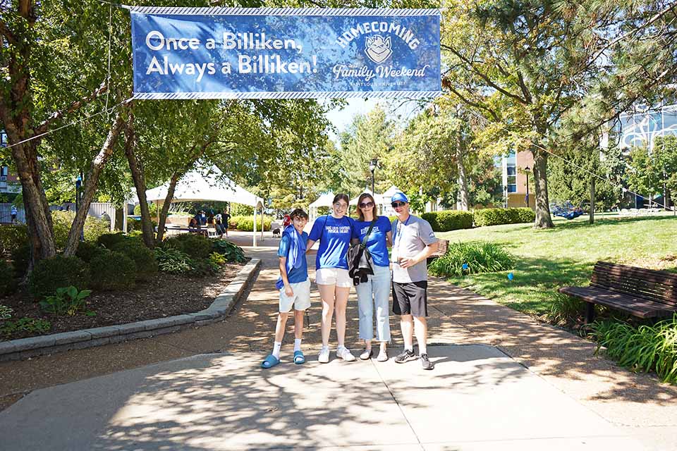 A family poses for a photo outside on campus under a sign that says Homecoming and Family Weekend.