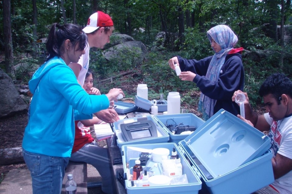 Students stand and sit at a picnic table, examining samples using biological equipment.
