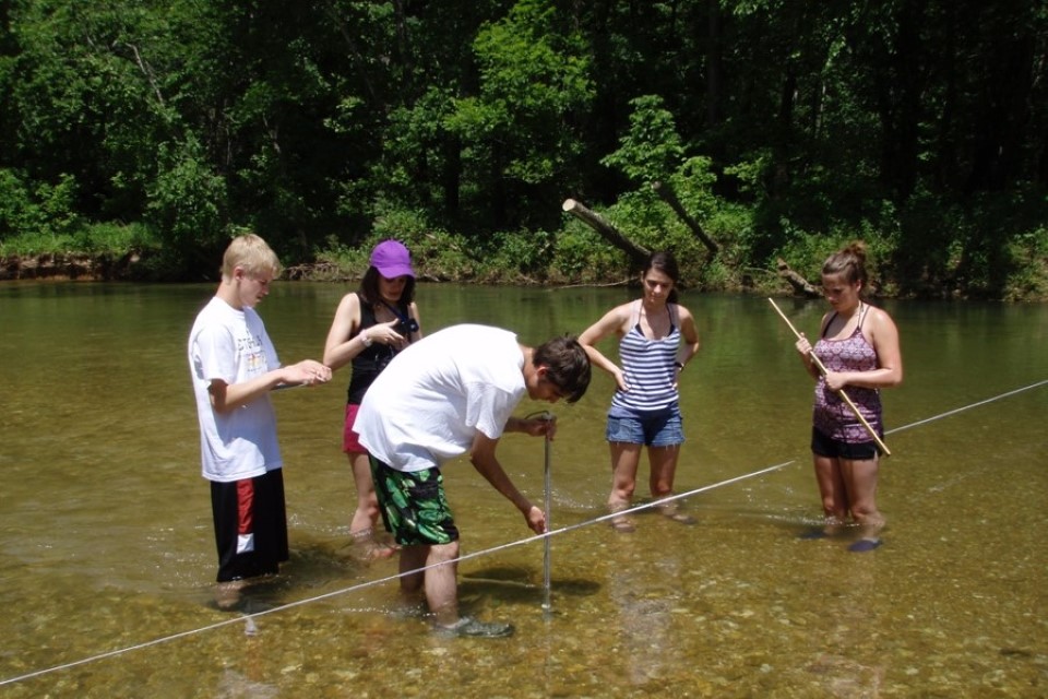 Students stand at knee level in a river, holding equipment and looking into the water.