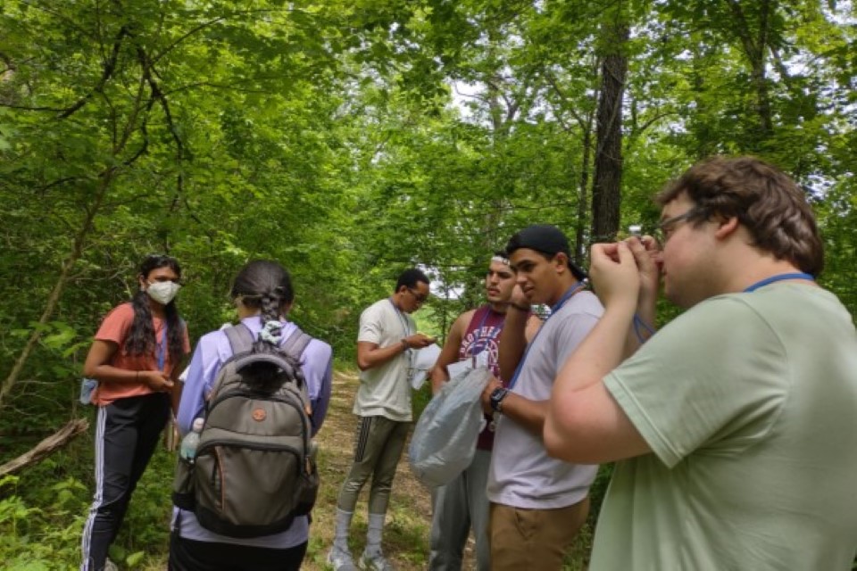 A group of students stands in a forest, examining papers and samples.