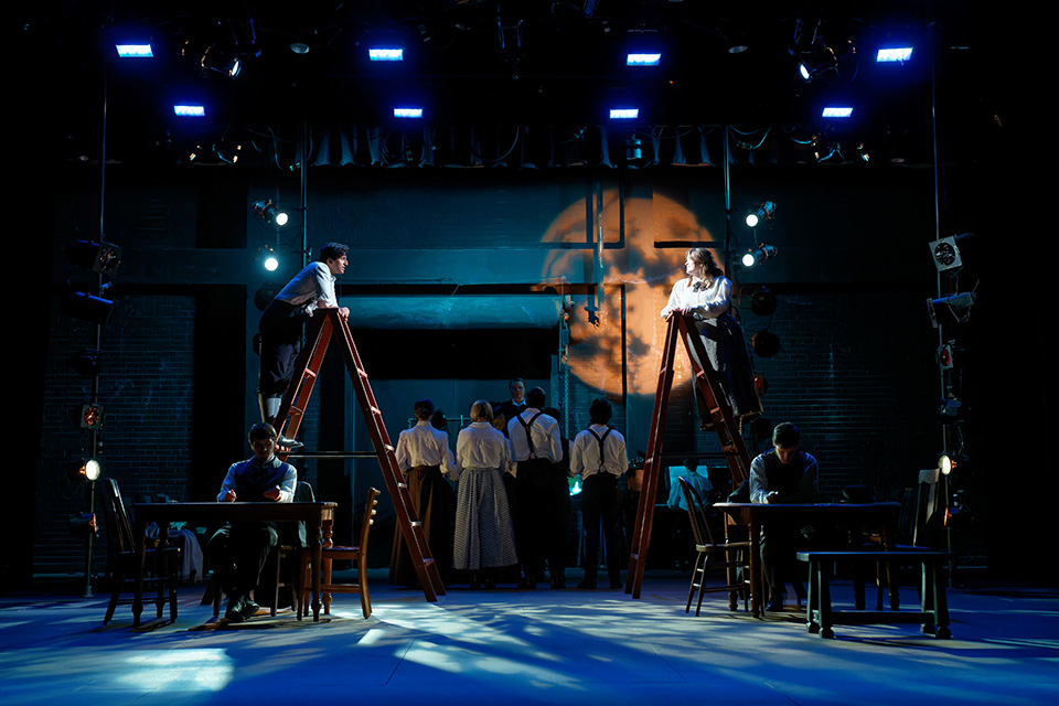 Two people stand on ladders with the moon overhead while other cast members stand with their backs to the audience and others sit at tables