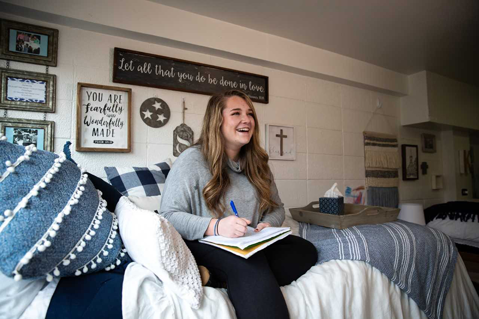 A student sits on her bed in a dorm room, laughing while writing in a notebook.