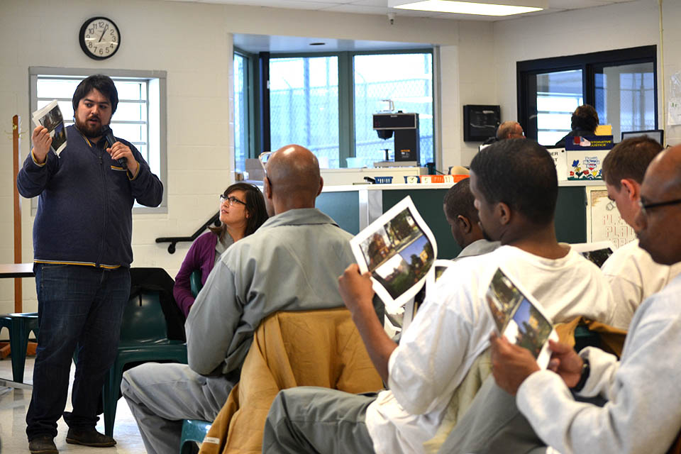 Students who are inmates at a correctional facility examine images during a lesson while a professor talks at the front of the room.