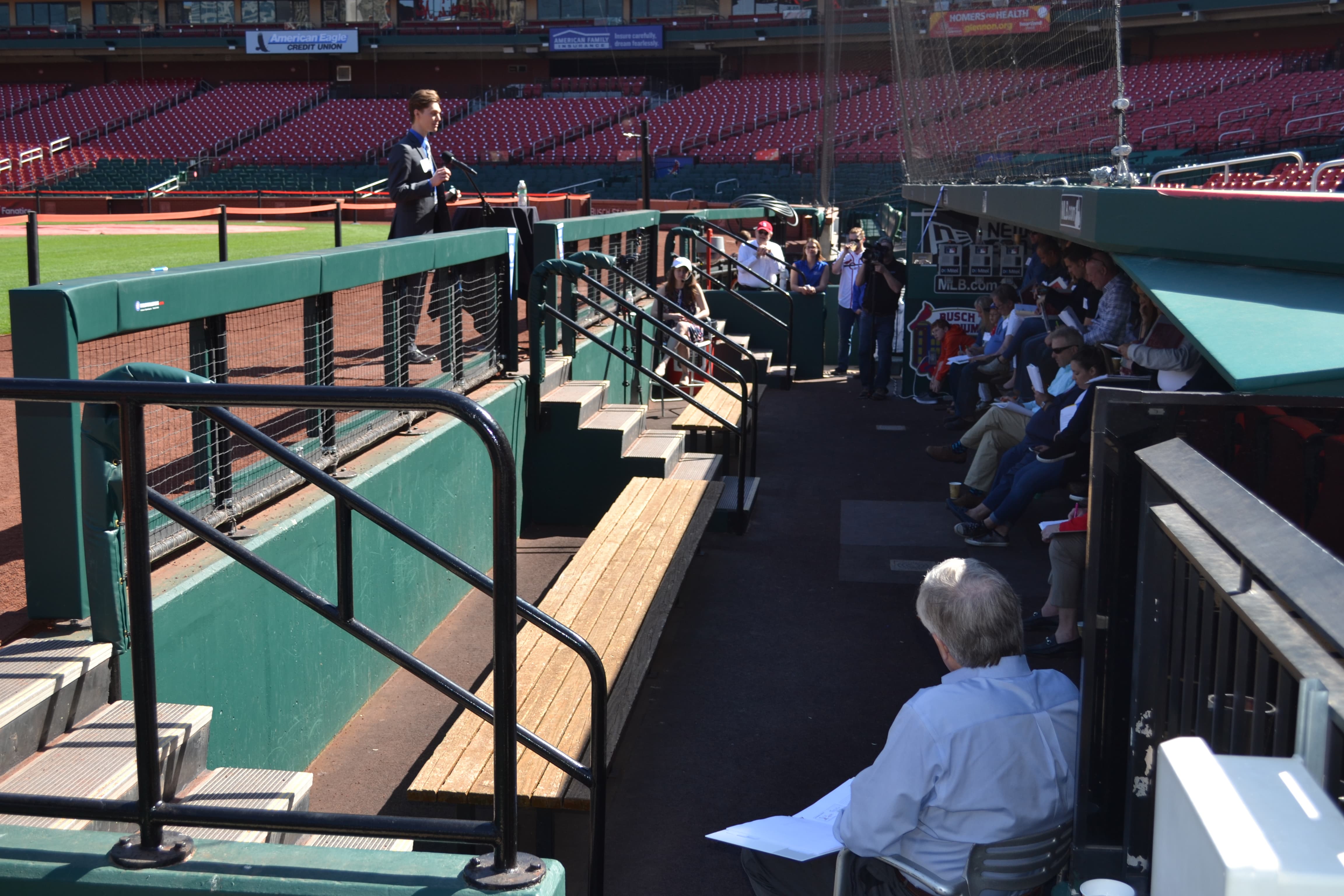 A student stands at the top of a dug out at Busch stadium speaking to a seated group 