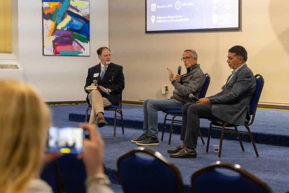 three men seated speaking on a stage in front of a crowd